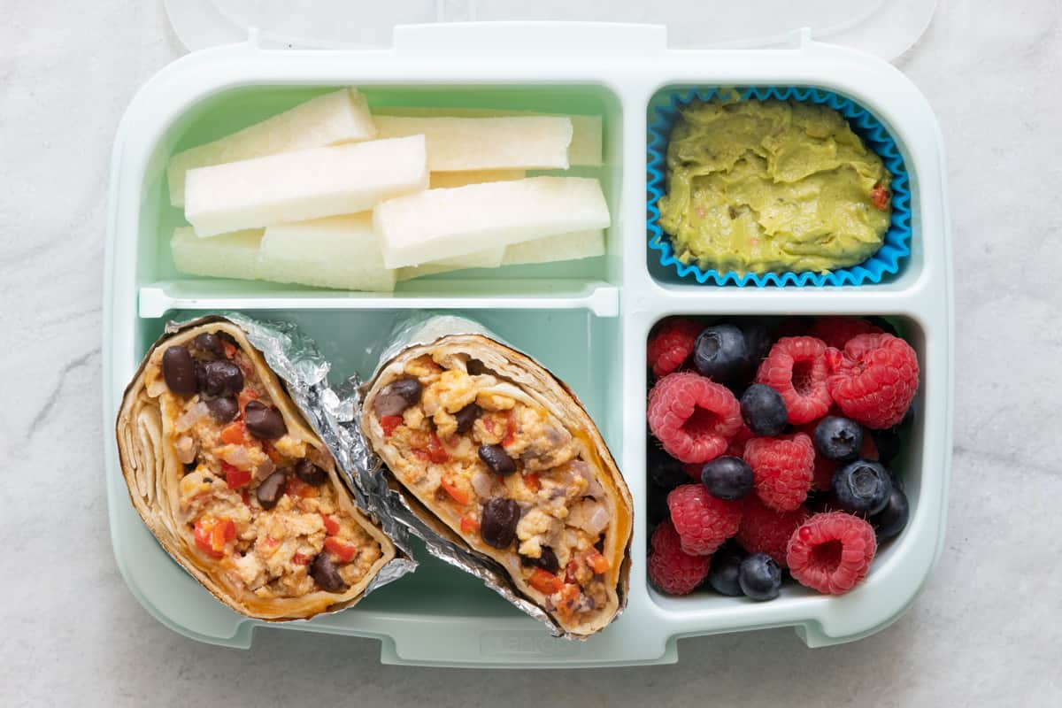 Lunchbox with 4 sections with different foods in each section: jicama sticks, breakfast burrito, guacamole, and fresh raspberries and blueberries.