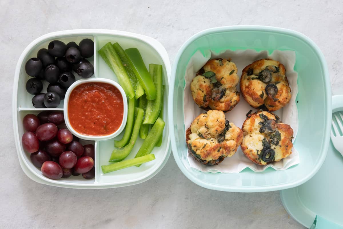 Stackable lunch container with individual sections and different foods in each: large container contains 4 bite sized pizzas on a napkin, and the sectioned container is filled with black olives, grapes, pizza sauce, and green pepper slices.