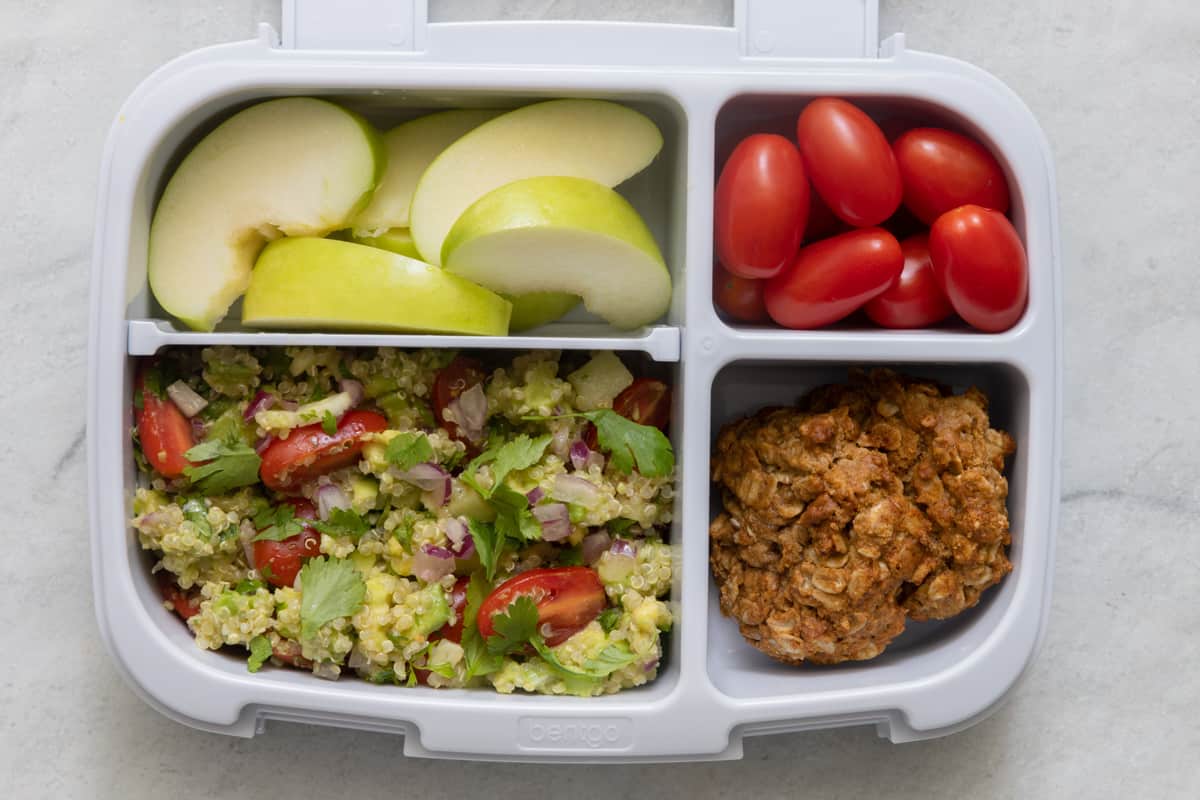 Lunchbox with 4 sections with different foods in each section: sliced green apples, qunioa salad, cherry tomatoes, and apple butter cookies.