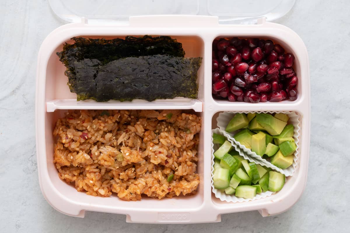 Lunchbox with 4 sections with different foods in each section: rice paper, tuna and rice mix, pomegranate seeds, and chopped cucumber and avocado in cupcake liner.