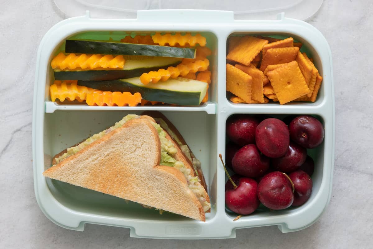 Lunchbox with 4 sections with different foods in each section: white bean avocado sandwich cut on a bias and stacked ontop of the other half, crinkle carrots and sliced cucumbers, cheese crackers, and fresh cherries.