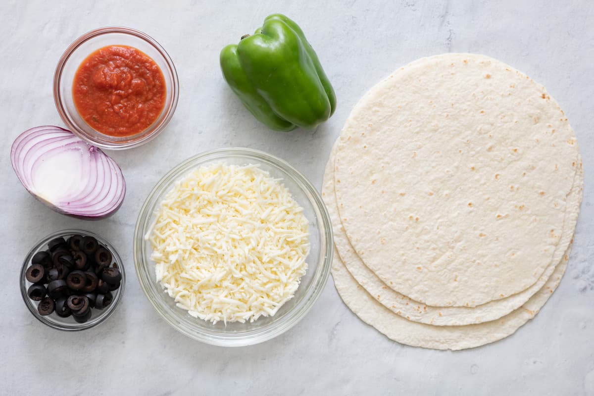 Ingredients for recipe before being prepped: marinara, half a red onion, sliced black olives, shredded cheese, green bell pepper, and tortilla shells.
