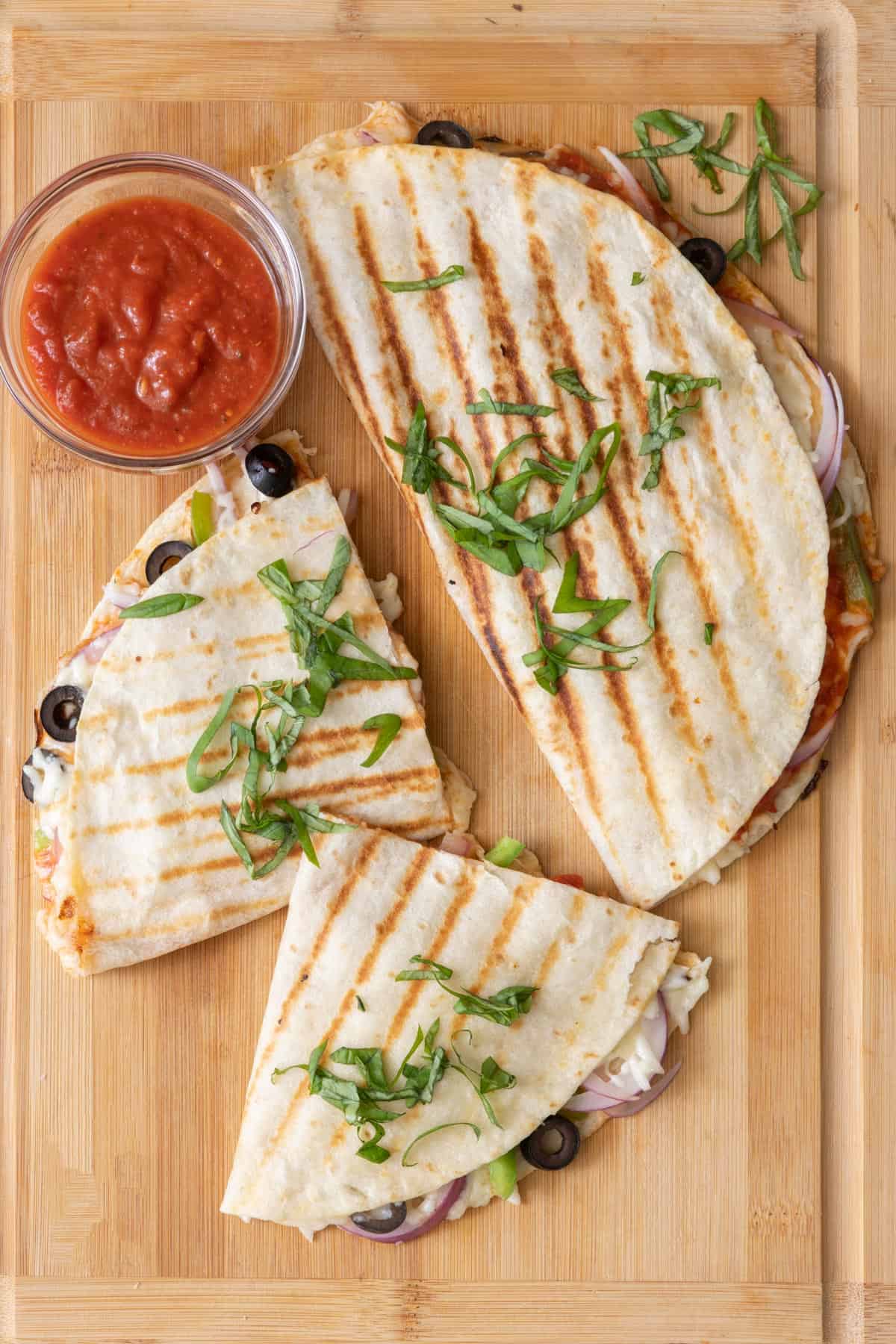 Pizza quesadillas on a cutting board with one cut in half next to a bowl of marinara sauce.