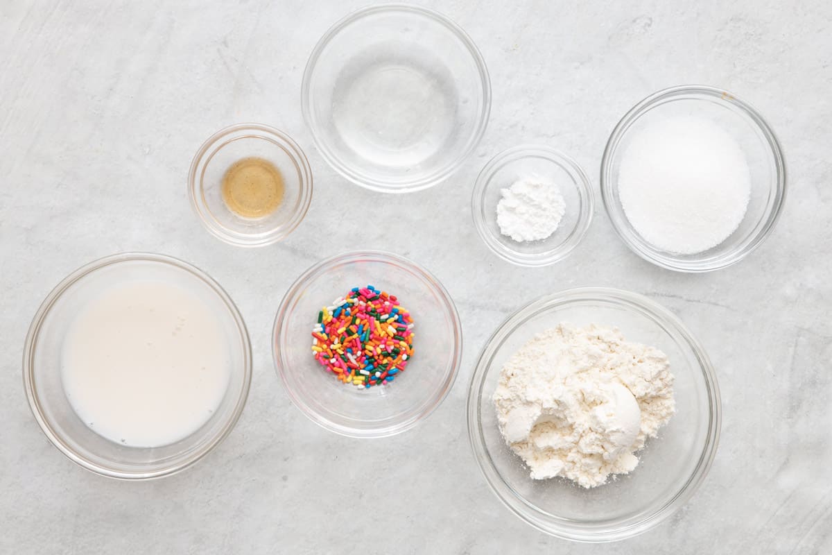 Ingredients in individual bowls: almond milk, vanilla extract, coconut oil, sprinkles, baking powder, sugar, and flour.