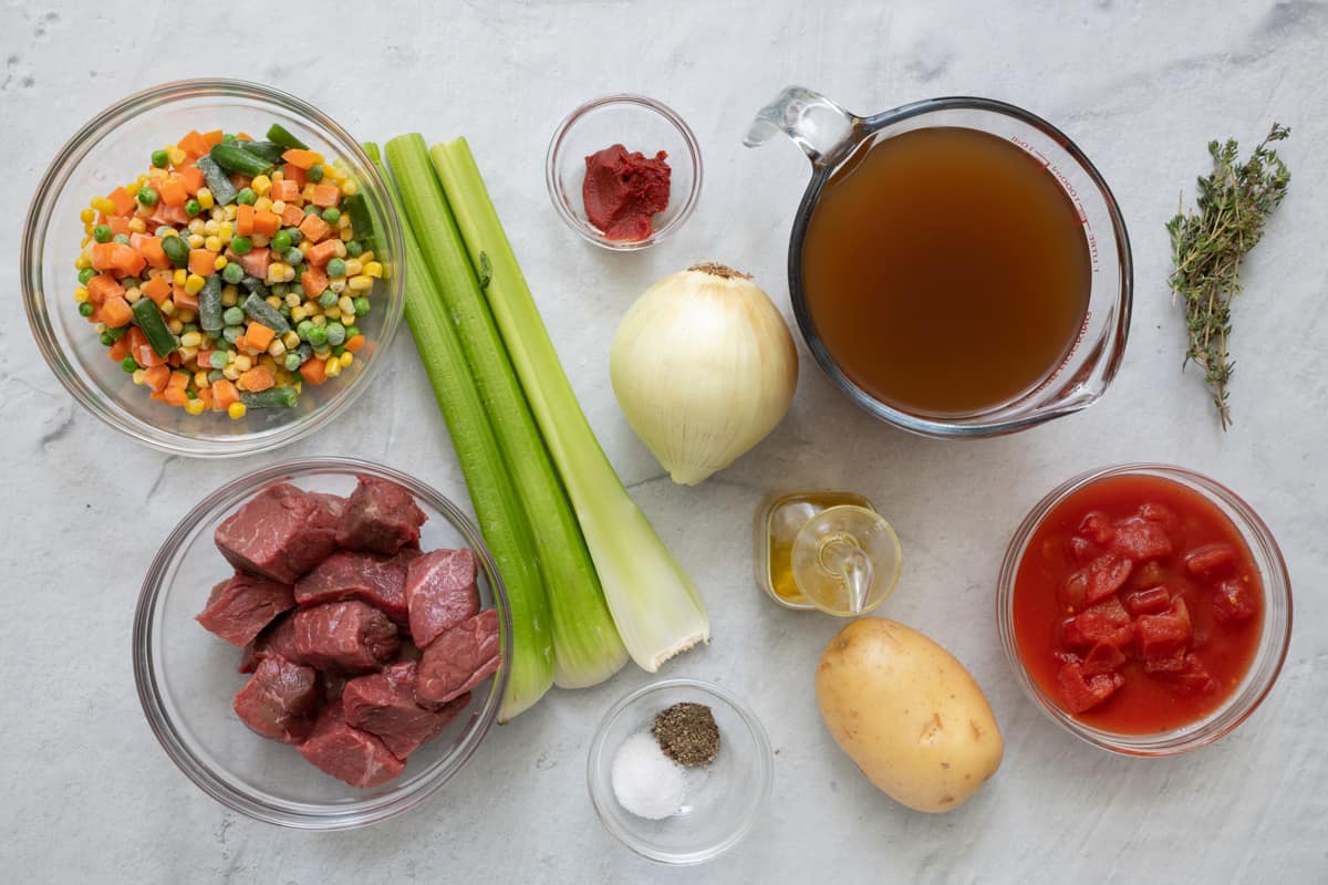 Ingredients for recipe in individual bowls and before being prepped: mixed frozen vegetables, chunks of beef, celery stalks, tomato paste, onion, oil, salt and pepper, broth, potato, and diced tomatoes.