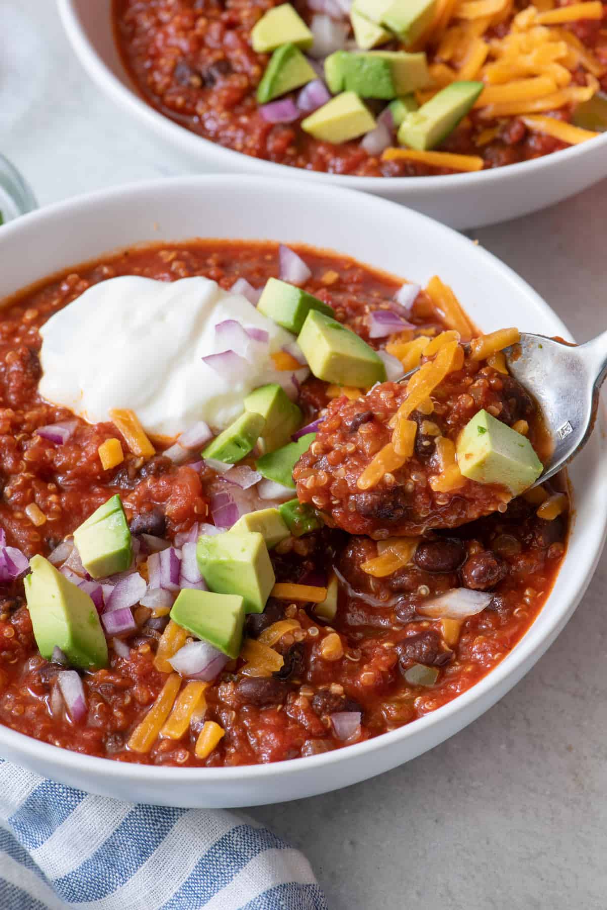 Two bowls of chili with one close up with a spoon scooping up some, garnished with greek yogurt, chopped avocado and red onions, and shredded cheese.