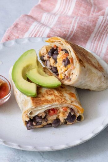 Burrito cut in half on a plate with slices of avocado on top and a small side of salsa.