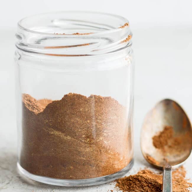 Small glass jar filled halfway with spice blend without a lid and a spoon dusted with pumpkin spice and some next to jar.