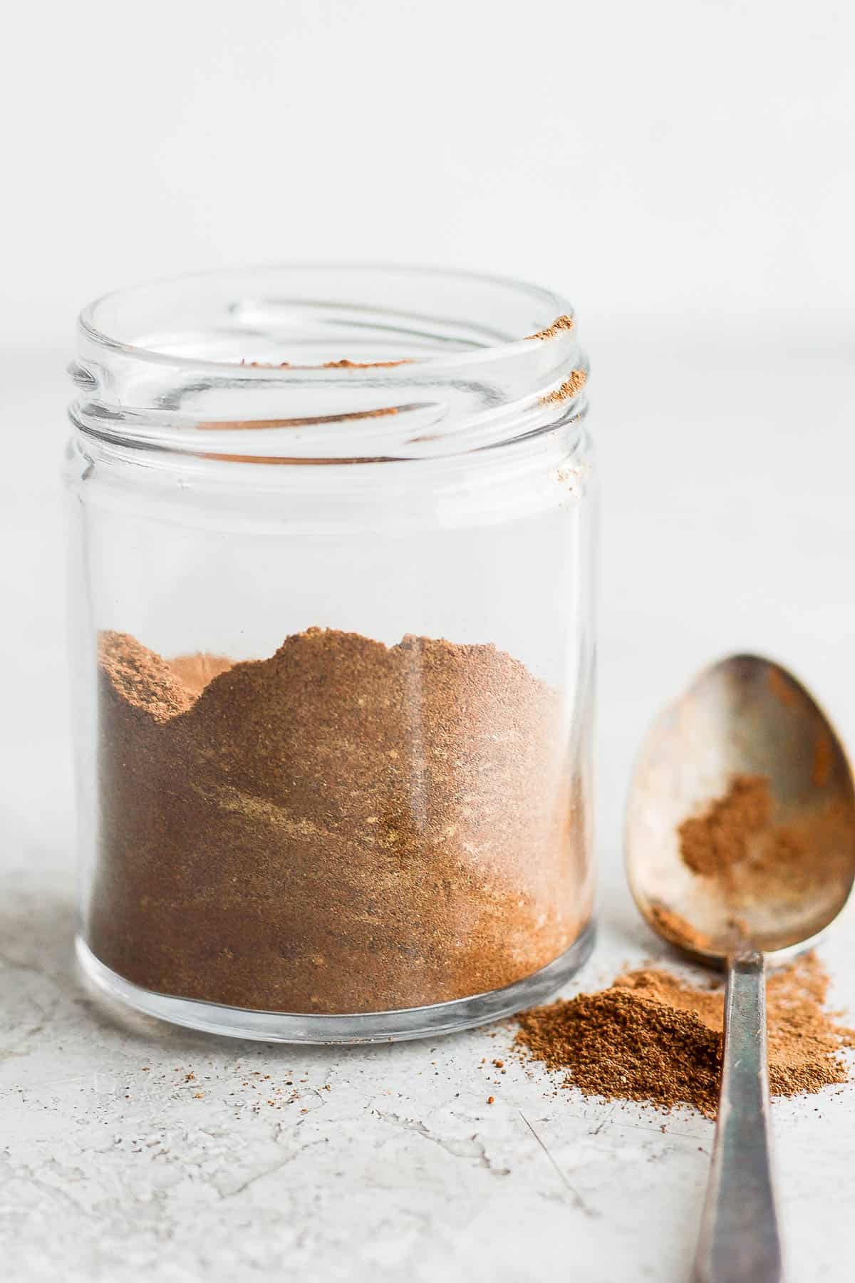 Small glass jar filled halfway with spice blend without a lid and a spoon dusted with pumpkin spice and some next to jar.