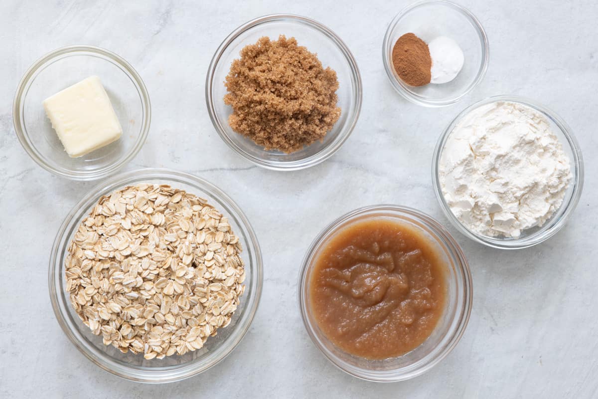 Ingredients for cookie recipe in individual small glass bowls: butter, oats, brown sugar, apple butter, baking soda and cinnamon, and flour.