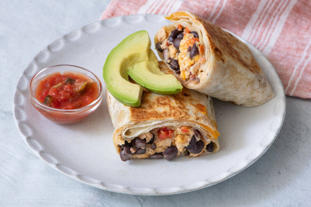 Burrito cut in half on a plate with slices of avocado on top and a small side of salsa.