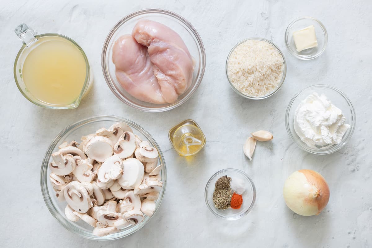 Ingredients for recipe in individual bowls: broth, sliced mushrooms, chicken breast, oil, spices, garlic cloves, rice, butter, sour cream, and onion.