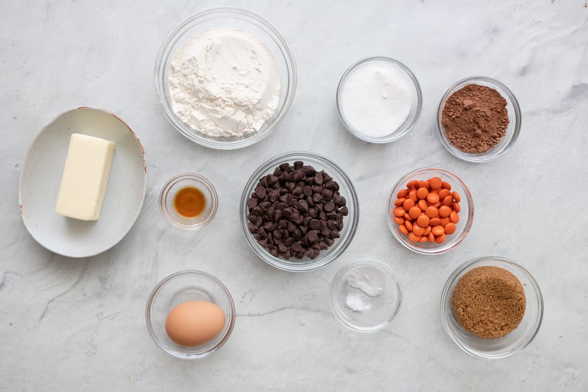 Ingredients for recipe measuring into individual dishes: butter stick, whlour, vanilla, egg, chocolate chips, sugar, baking powder, orange candies, cocoa powder, and brown sugar.