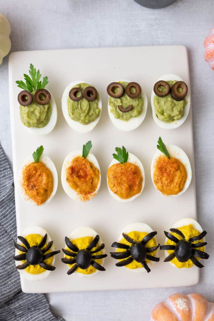 White board with halloween decorated devilled eggs. Top row avocado with googly eggs made of olives, second row decorated to look like a pumpkins, and third row yellow with spiders made of black olives.