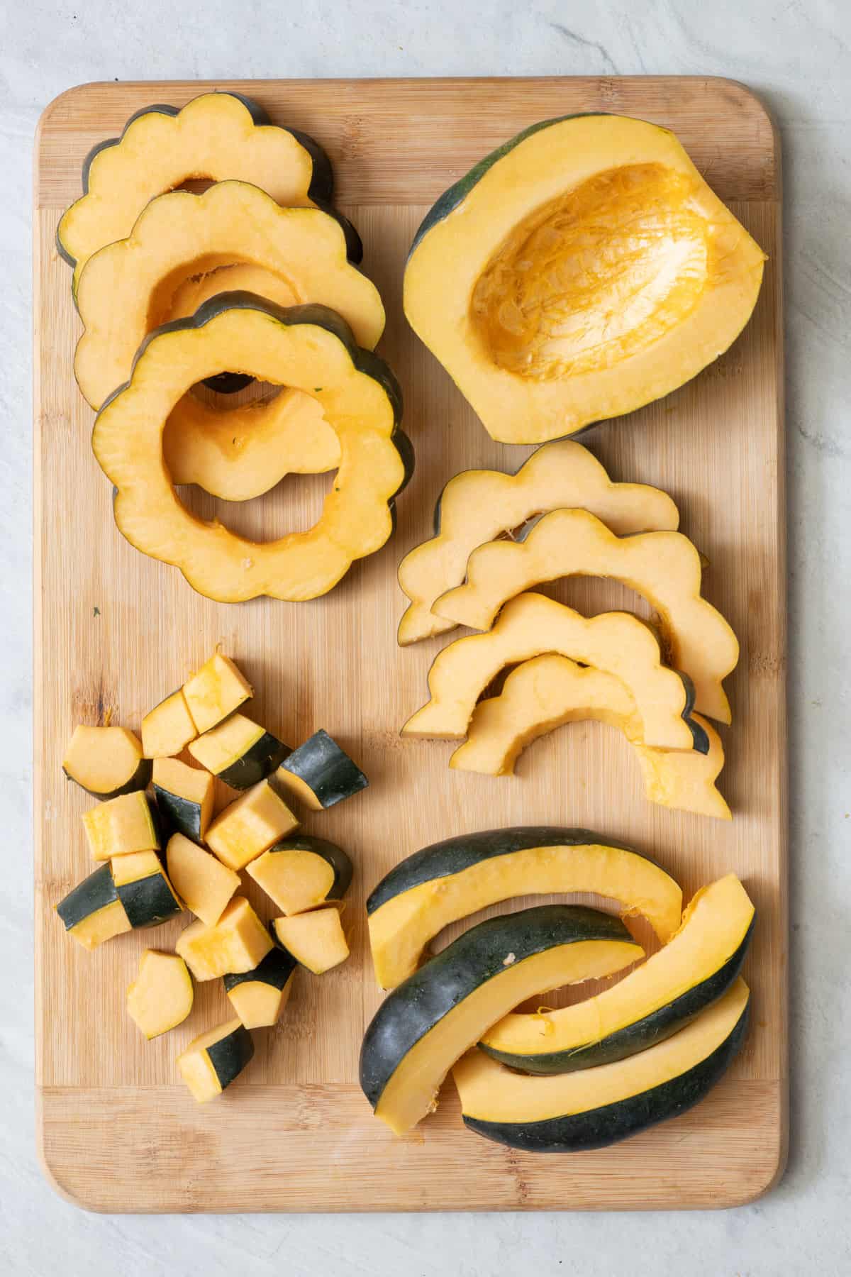 Cutting board with one half, few slices, few wedges, cubes, and few circles of acorn squash.