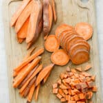 A cutting board with sweet potatoes cut 4 different ways: wedges, slices, sticks, and cubes.