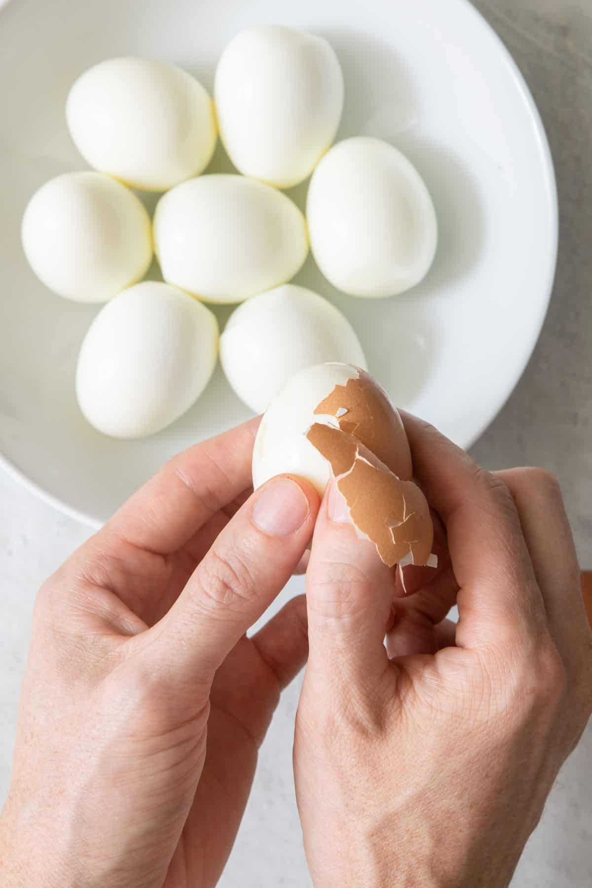 Hands peeling a brown egg with a plate of peeled eggs below.