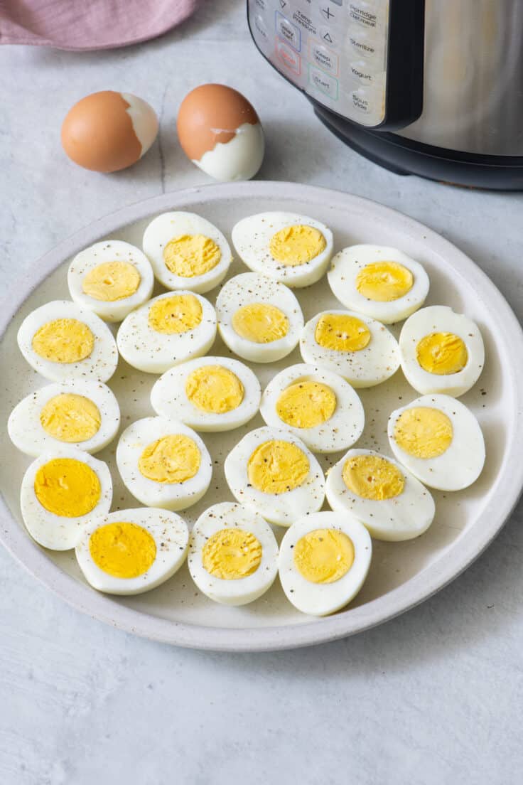 A plate of hard cooked eggs cut in half with salt and pepper. Two half peel eggs sit behind plate next to a half shown instant pot.