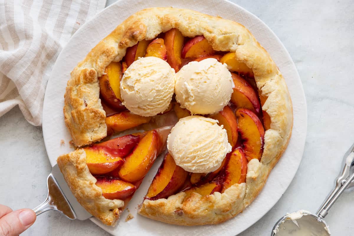 Galette filled with peaches with a slice being removed and topped with 3 scoops of vanilla ice cream.