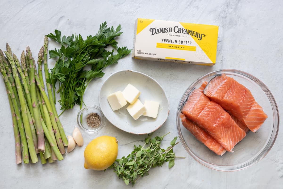 Ingredients for recipe before being prepped: fresh asparagus, oregano, and parsley, garlic, Danish Creamery butter sticks, seasonings, a whole lemon, and 4 salmon fillets with skin on in a bowl.