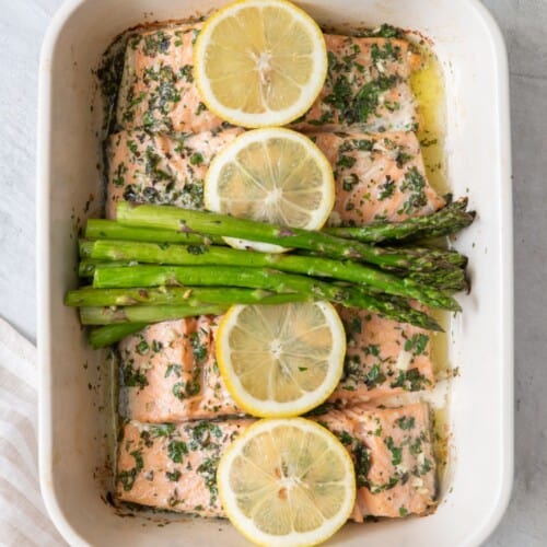 Roasted salmon and asparagus in a baking dish with lemons resting over it.