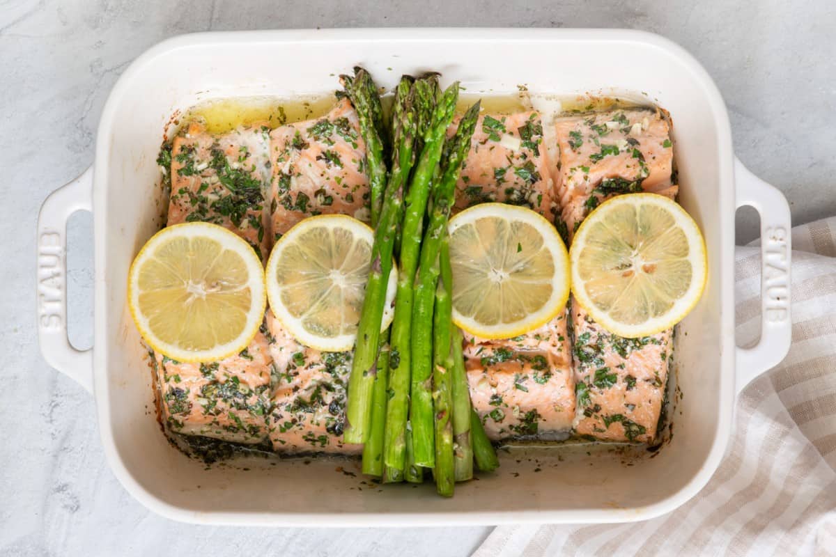 Roasted salmon and asparagus in a baking dish with lemons resting over it.