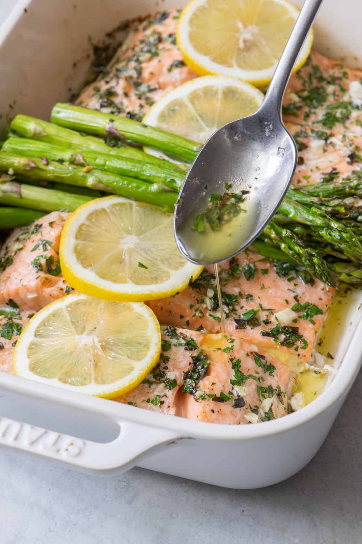 Four salmon filets roasted in a baking dish topped with roasted asparagas and fresh lemon slices and a spoon pouring sauce over them.