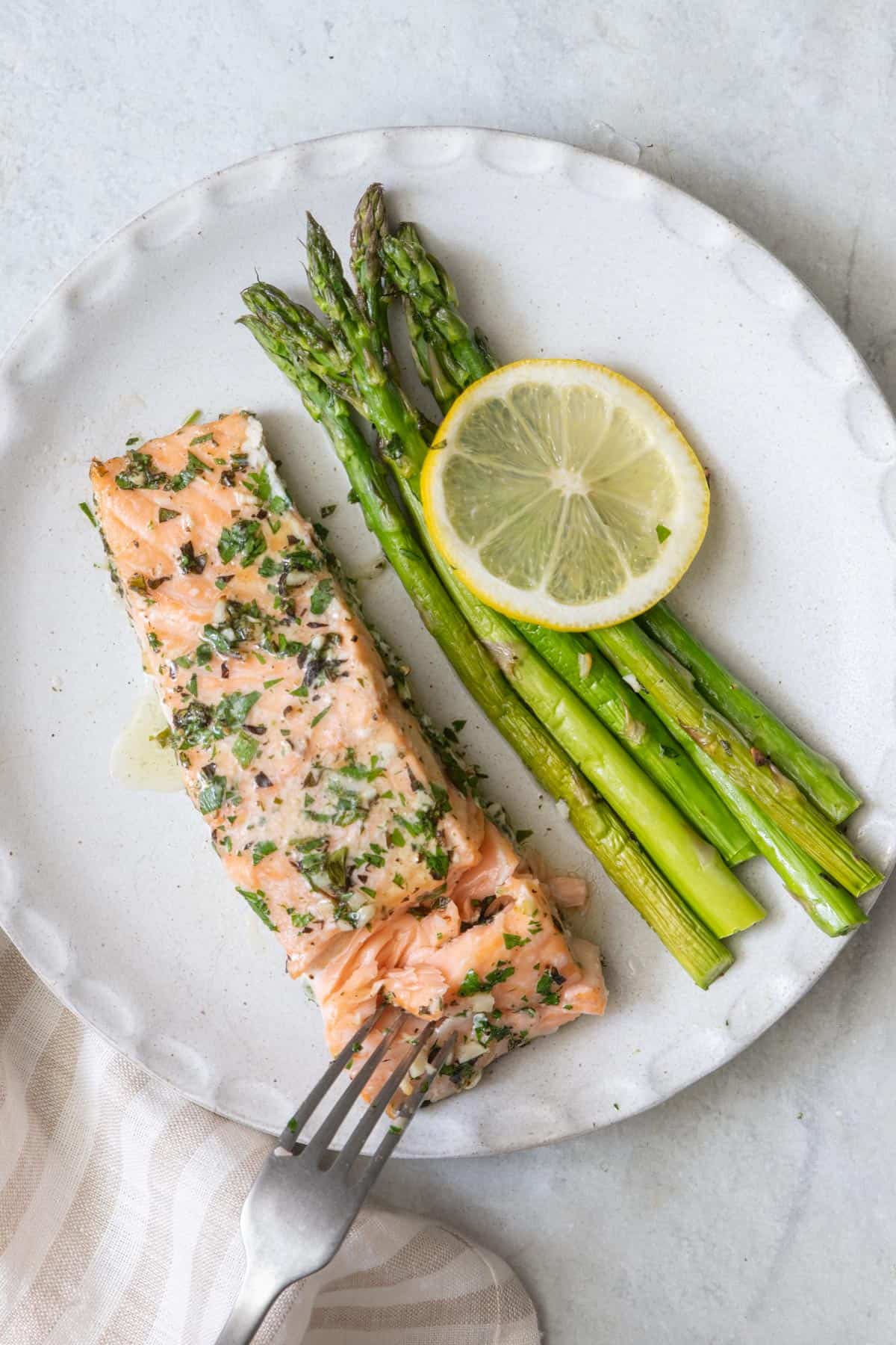 Roasted salmon, asparagus, and a fresh lemon slice on a white plate and a fork flaking away some of the fish.