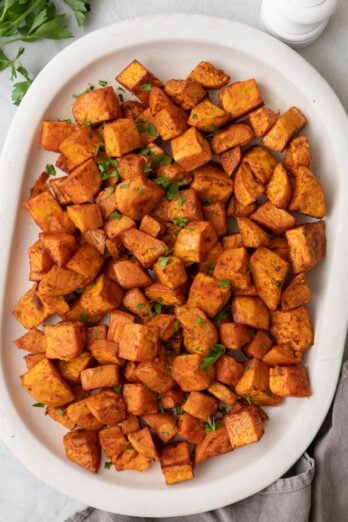 Diced roasted sweet potatoes on large white oval serving dish garnished with fresh parsley.