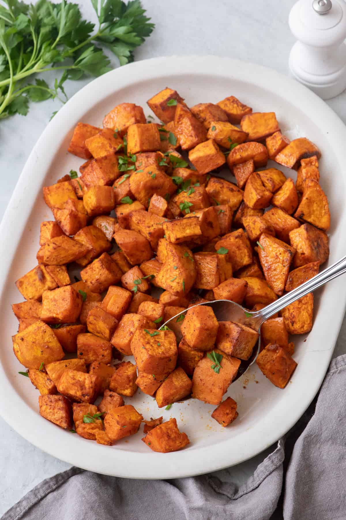 Diced roasted sweet potatoes on large white oval serving dish garnished with fresh parsley and a spoon scooping some.