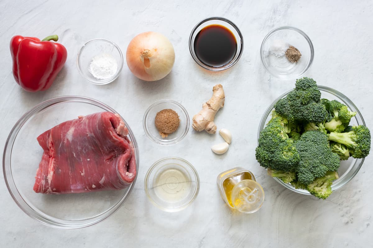 Ingredients for recipe before being prepped: whole red pepper, flank steak folded in a bowl, cornstarch, whole onion, spices, rice vinegar, soy sauce, piece of fresh ginger, garlic cloves, salt and pepper, oil, and a bowl of fresh brocolli.