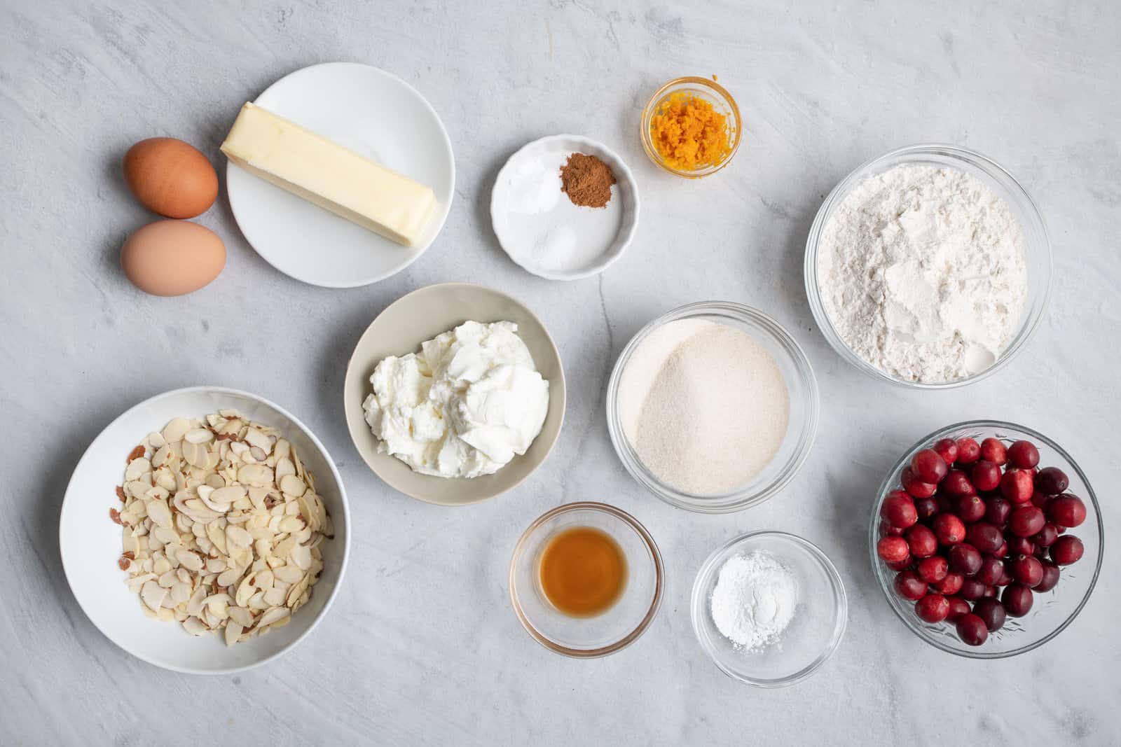 Ingredients for recipe in individual bowls: 2 eggs, sliced almonds, butter stick, greek yogurt, spices, orange zest, vanilla, sugar, baking powder, flour, and fresh cranberries.