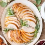 Overhead shot of sliced turkey breast on a large platter with fresh herbs with sweet potatoes and cranberry sauce nearby.