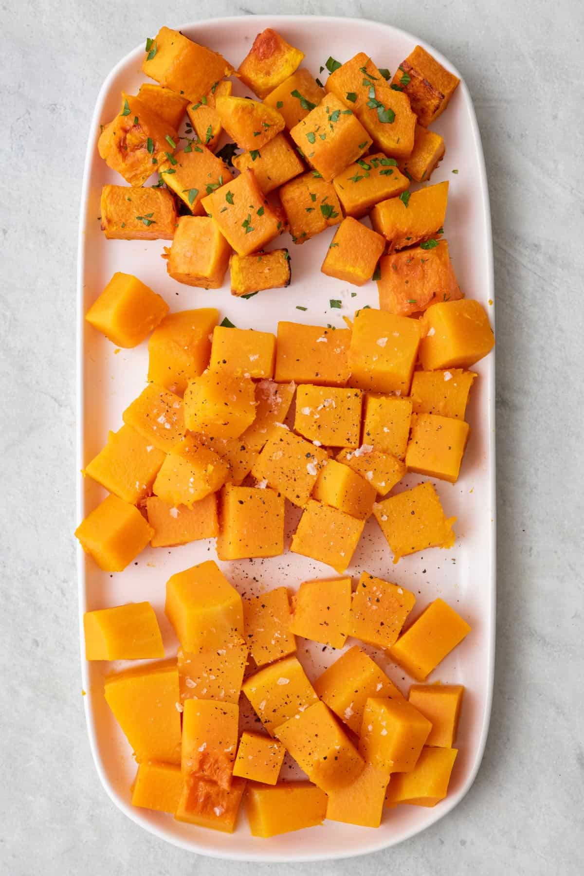 Butternut squash cut into cubes and cooked three different ways on a large platter, some garnished with pasley and flaky salt.