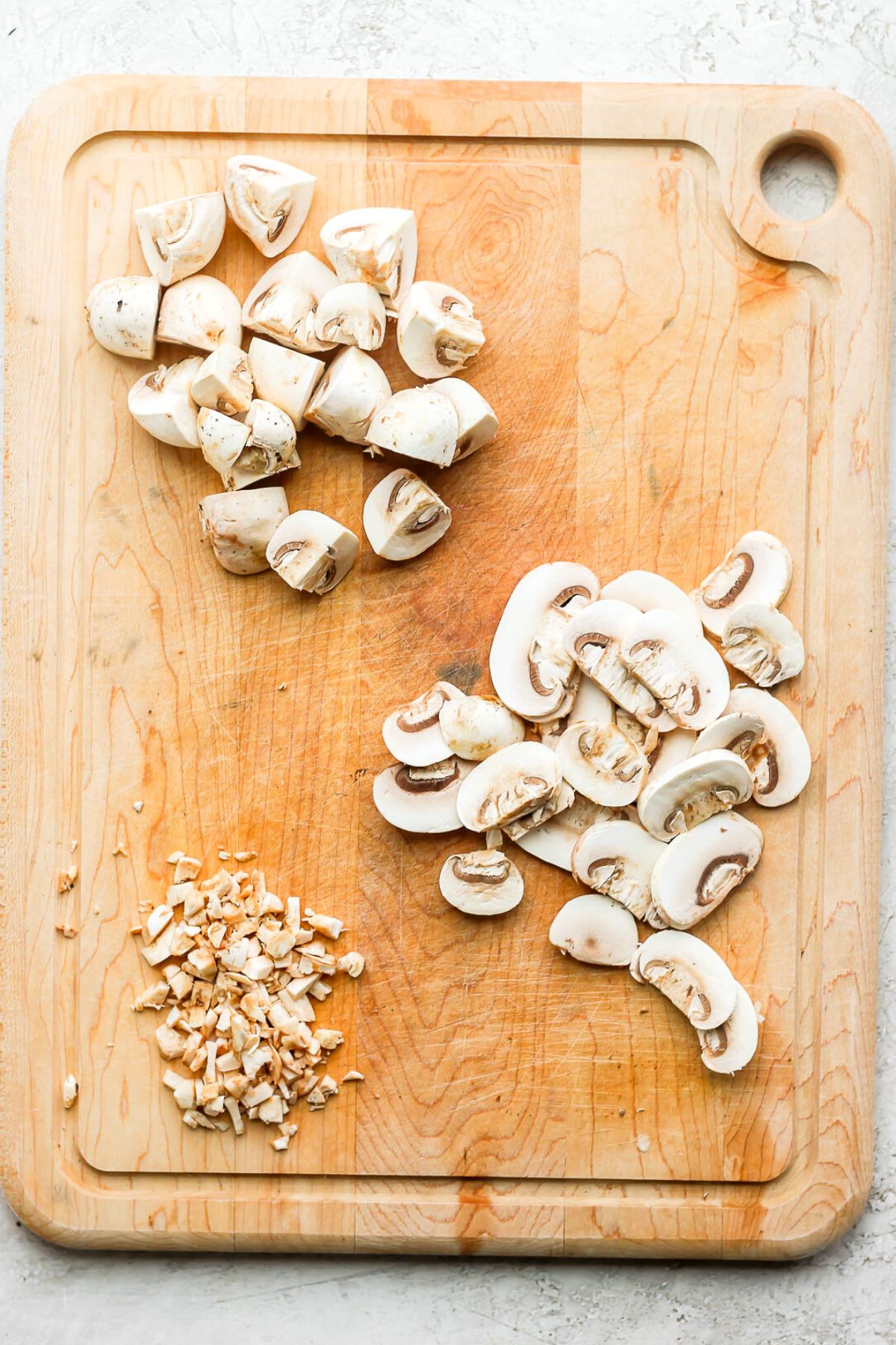 Cutting board with 3 different types of cut mushrooms: quartered, sliced, and minced.