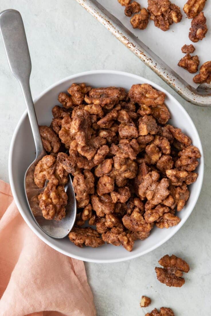 Candied walnuts in a small bowl with a spoon dipped in and baking sheet with more on it nearby.