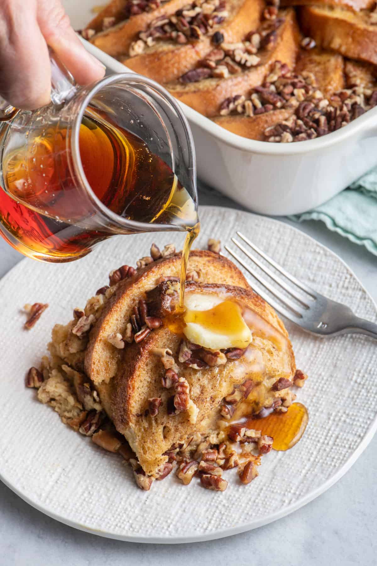 Serving of baked overnight french toast on a textured white plate with maple syrup being poured over and casserole dish of recipe in background.