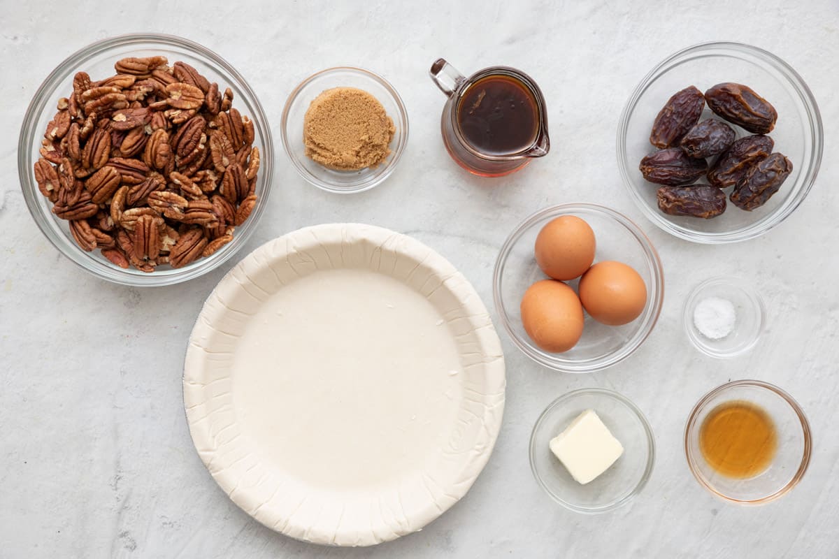 Ingredients for recipe in individual bowls before prepped: pecan halves, brown sugar, frozen pie shell in pan, maple syrup, 3 eggs, butter, dates, salt, and vanilla.