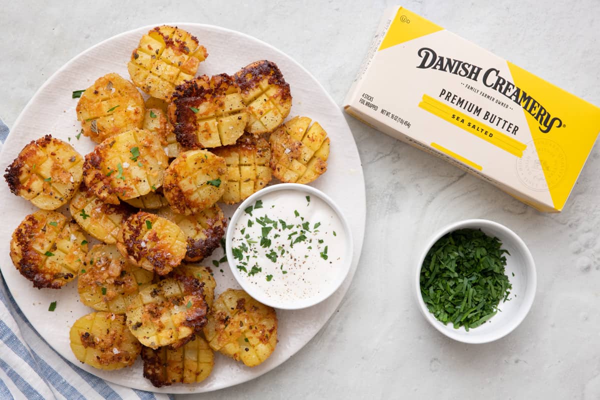 Plate of roasted parmesan potatoes with a small pinch bowl of ranch on the plate garnished with fresh chopped parsley, more chopped parsley in a small bowl nearby, and a box of Sea Salted Danish Creamery Premium Butter.