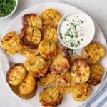 Roasted parmesan potatoes on a serving dish cut side up to show crosshatch and crispy parmesan with a small pinch bowl of ranch, garnished with fresh parsley and serving spoon sitting on plate.