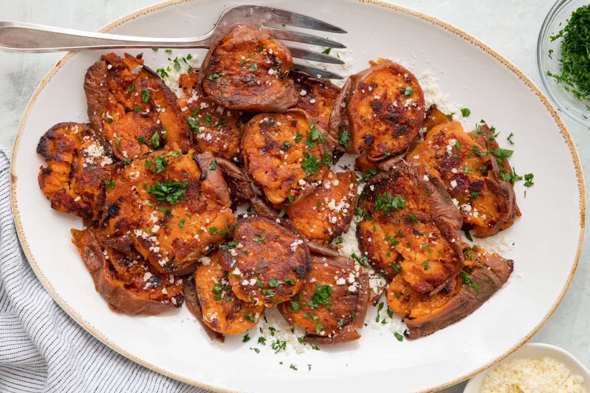 Smashed sweet potato rounds on a large oval platter with a fork set inside and garnished with fresh parsley and parmesan.