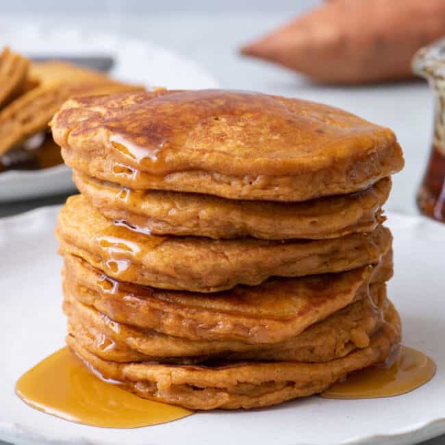 Side view of 6 sweet potato pancakes stacked on a white plate with maple syrup poured on top.