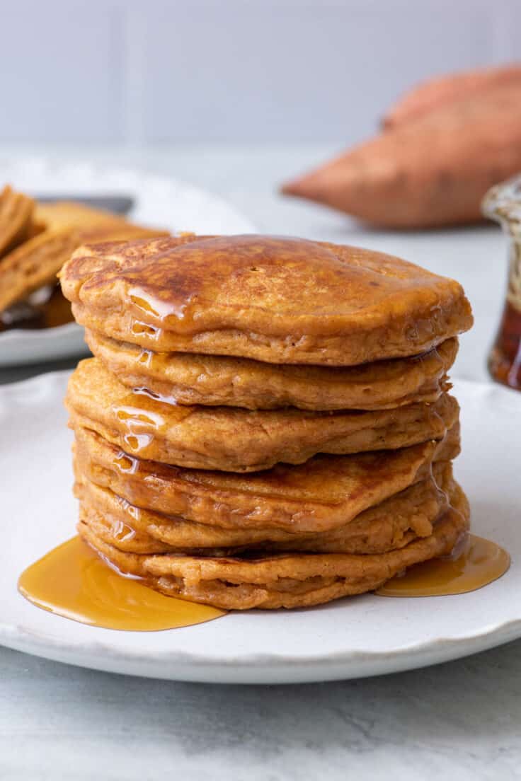 Side view of 6 sweet potato pancakes stacked on a white plate with maple syrup poured on top.