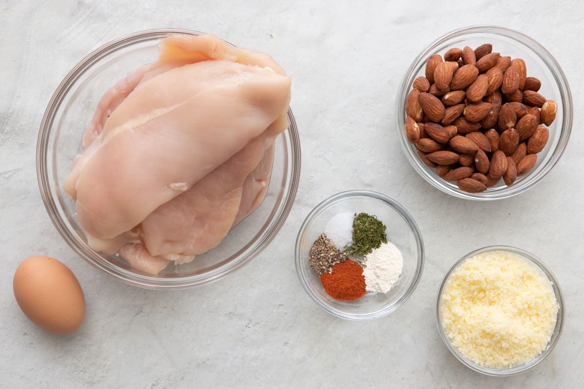 Ingredients for recipe in individual bowls before prepped: chicken breast, egg, seasonings, whole almonds, and parmesan cheese.