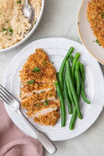 Small plate with almond crusted chicken partially sliced to show texture and garnished with fresh parsley served with fresh green beans with a bowl of risotto nearby.