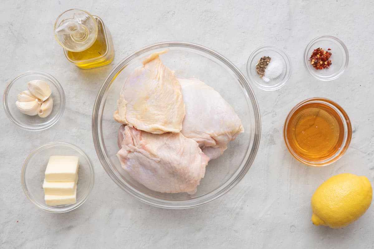 Ingredients for recipe in individual bowls: oil, garlic cloves, butter, bone in skin on chicken thighs, salt and pepper, red pepper flakes, honey, and a lemon.