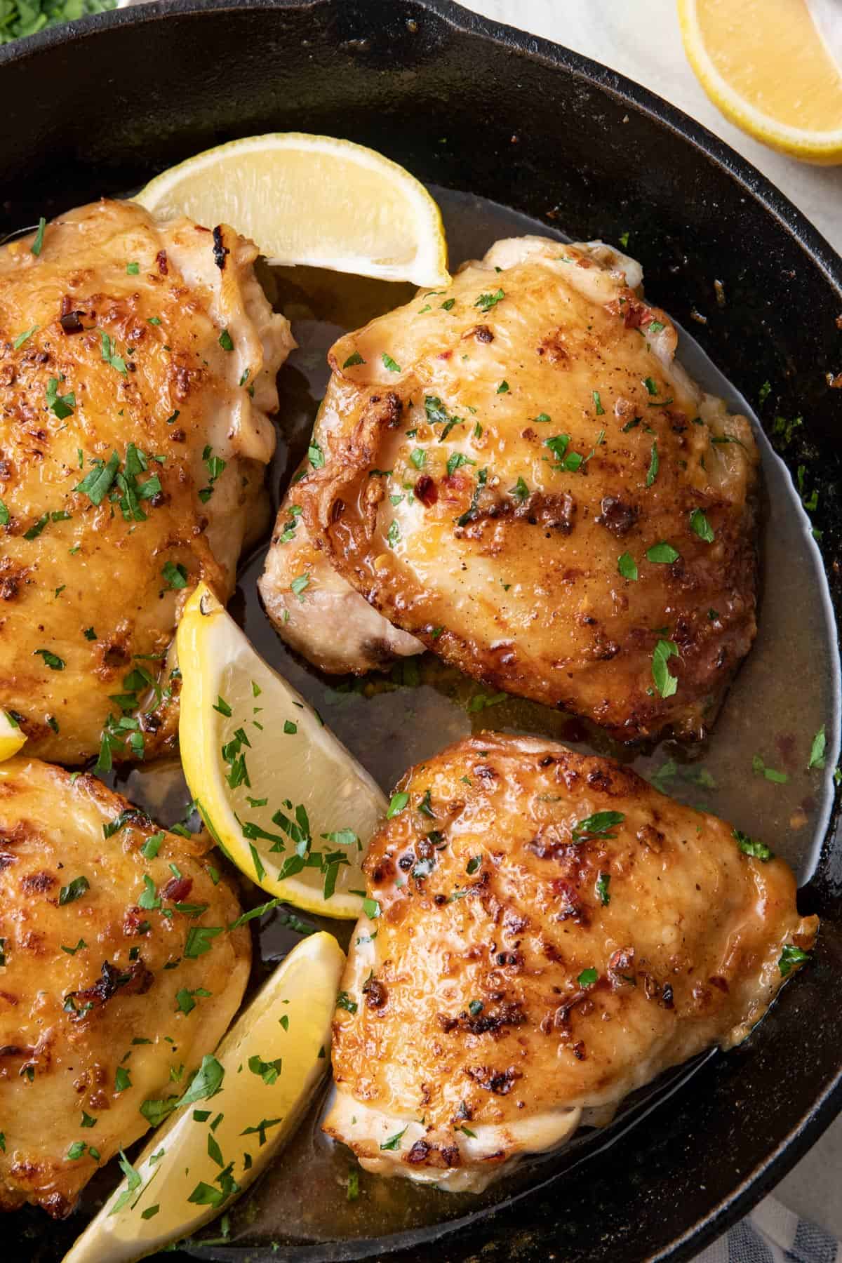 Close up of oven roasted chicken thighs with browned butter honey sauce garnished with lemon wedges and fresh herbs in a cast iron skillet.