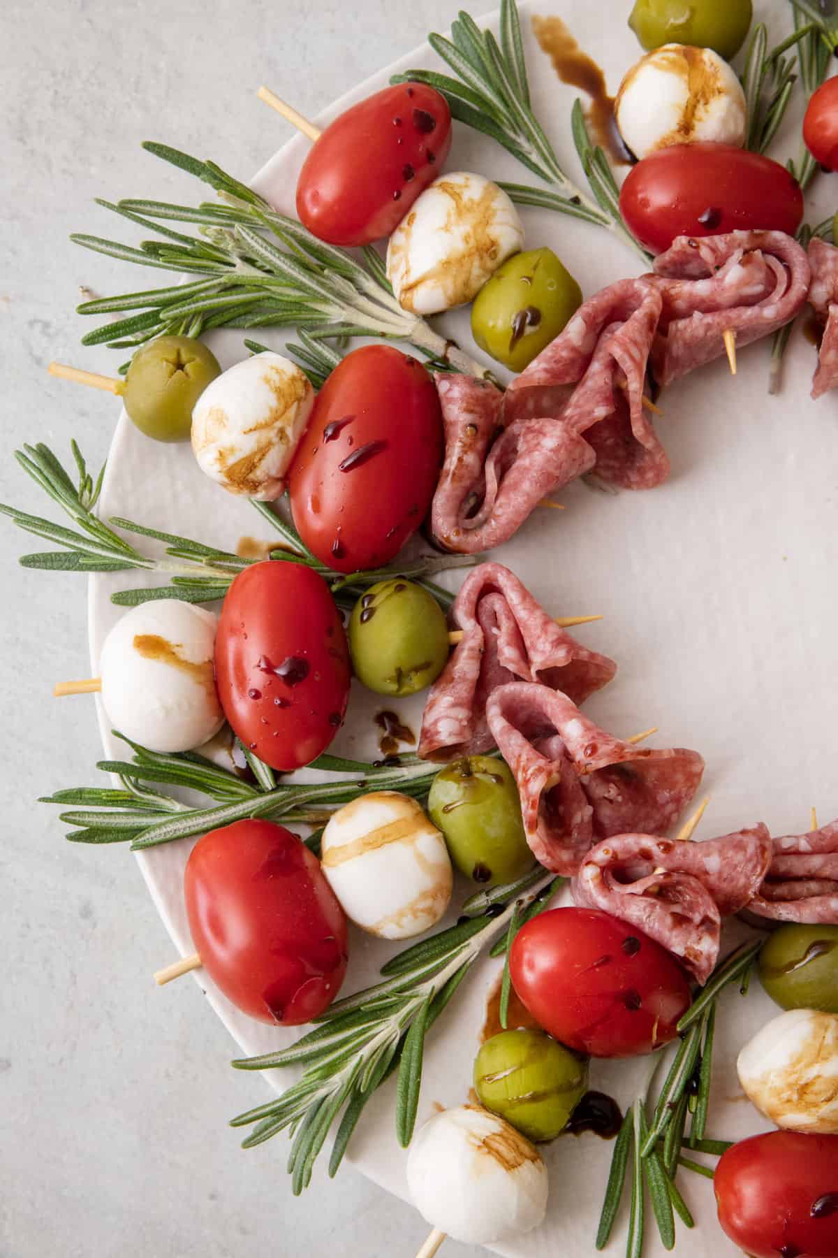 Close up of charcuterie wreath show each toothpick with an olive, mozzerella ball, tomato, and salami with rosemary between each and drizzled with balsamic glaze.