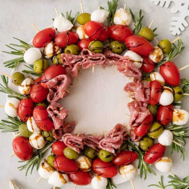Charcuterie wreath full of individual skewers of tomatoes, salami, green olive, and mozzerella drizzled with balsamic glaze, garnished with rosemary sprigs and surrounded by white christmas decor.
