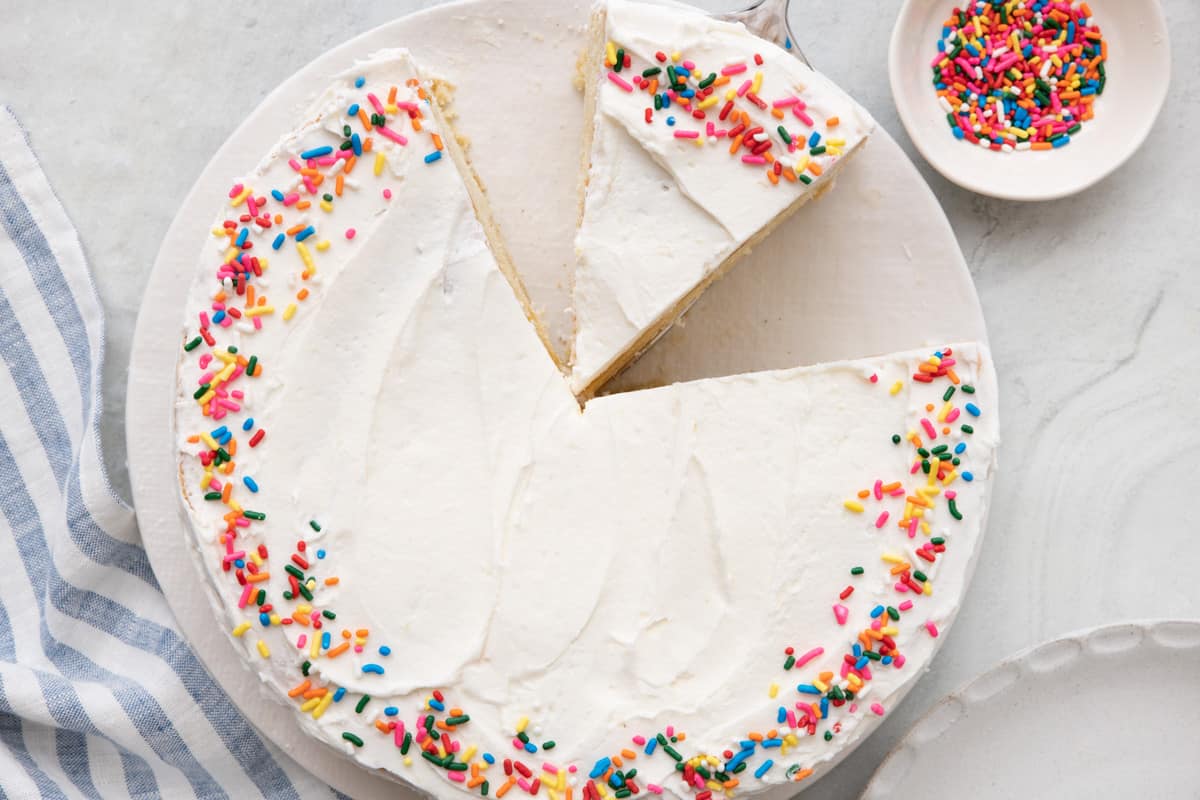 Overhead shot of vanilla cake with sprinkles around edge, a few pieces cut out with one slightly moved from remaining cake, and a small dish of sprinkles nearby.